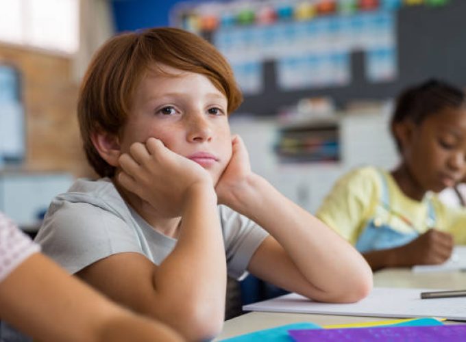 Tired school boy with hand on face sitting at desk in classroom. Bored schoolchild sitting at desk with classmates in classroom. Frustrated and thoughtful young child sitting and looking up.